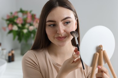Photo of Beautiful woman drawing freckles with brush in front of little mirror indoors