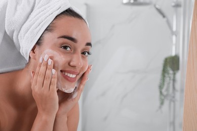Beautiful young woman applying cleansing foam onto face in bathroom, space for text. Skin care cosmetic