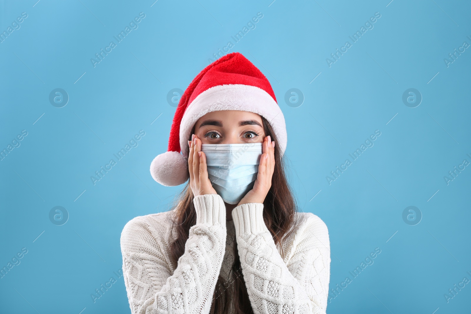 Photo of Beautiful emotional woman wearing Santa Claus hat and medical mask on light blue background