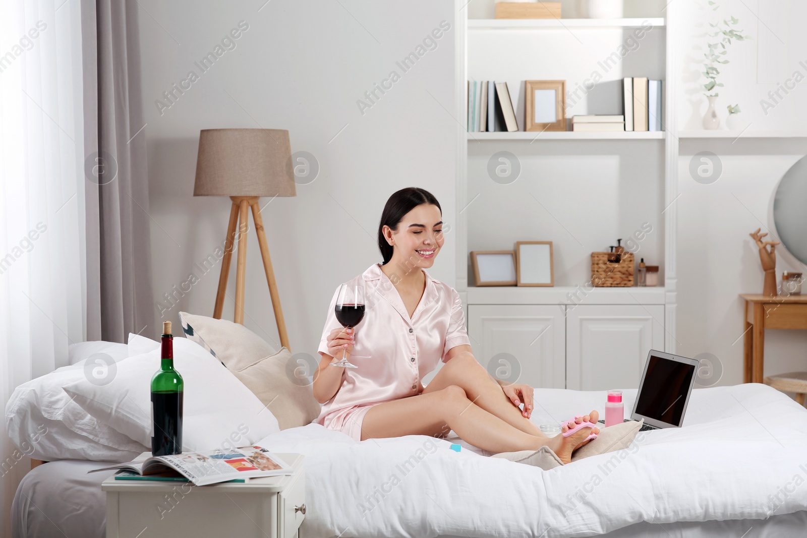 Photo of Beautiful young woman with glass of wine giving herself pedicure on bed at home