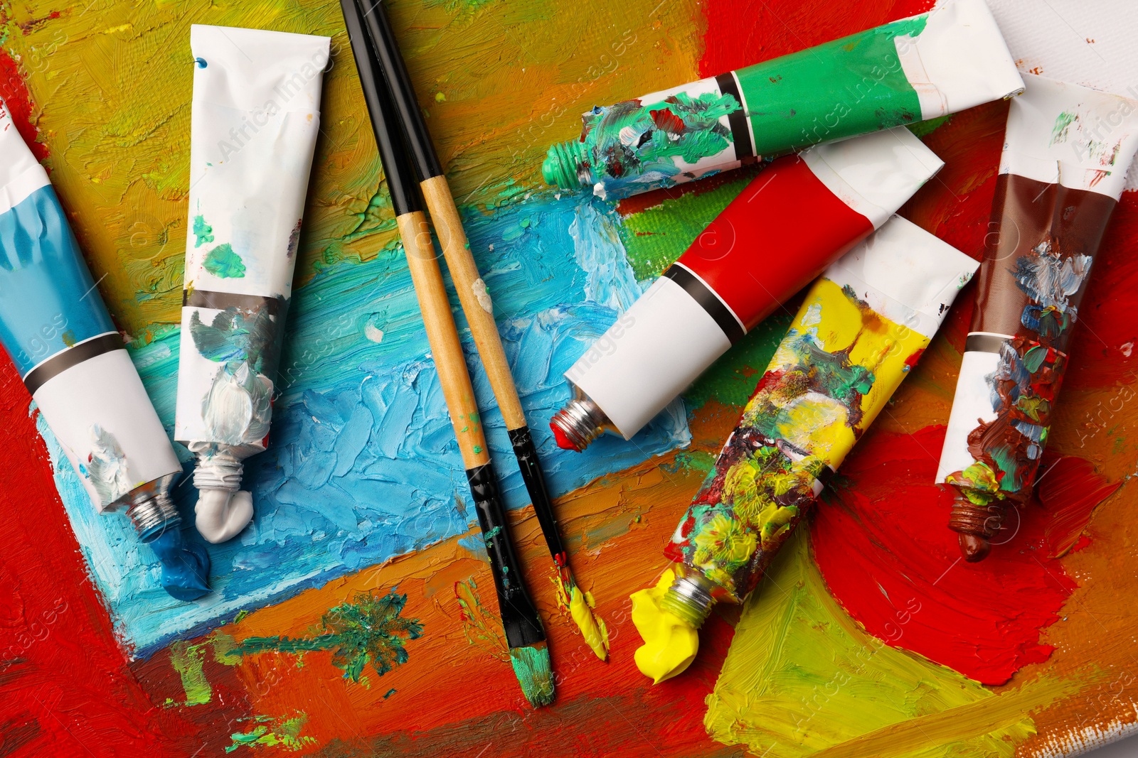 Photo of Tubes of colorful oil paints and brushes on canvas with abstract painting, flat lay