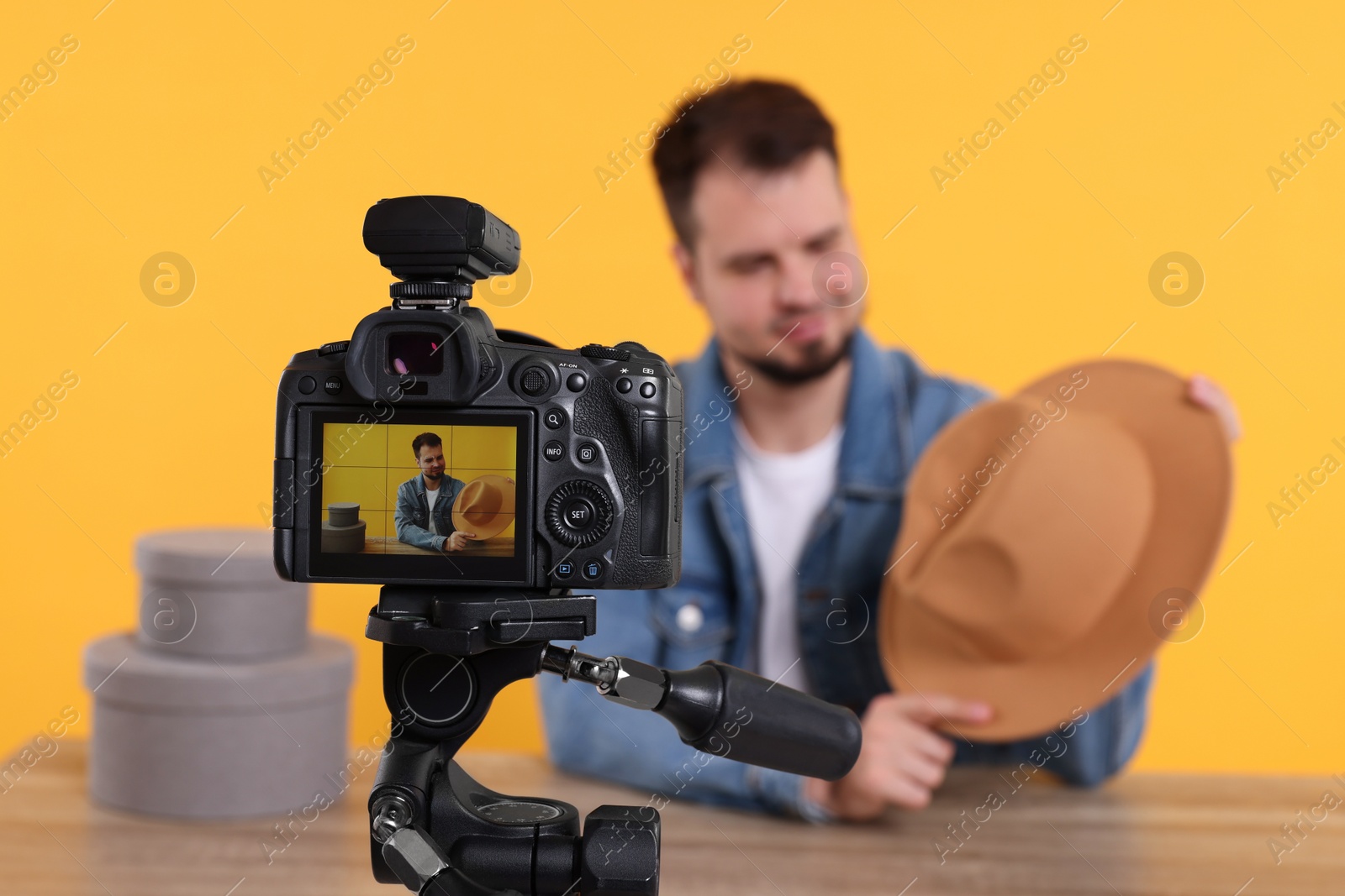 Photo of Fashion blogger showing hat while recording video at table against orange background, focus on camera