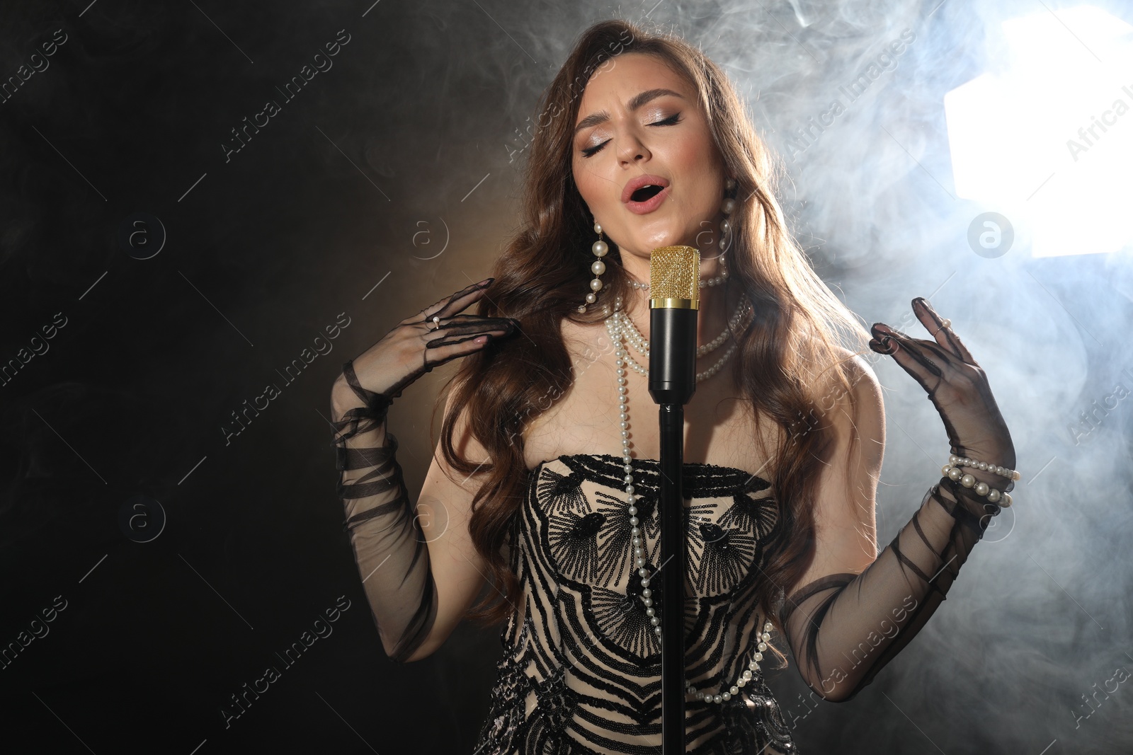 Photo of Beautiful young woman with microphone singing on dark background with smoke