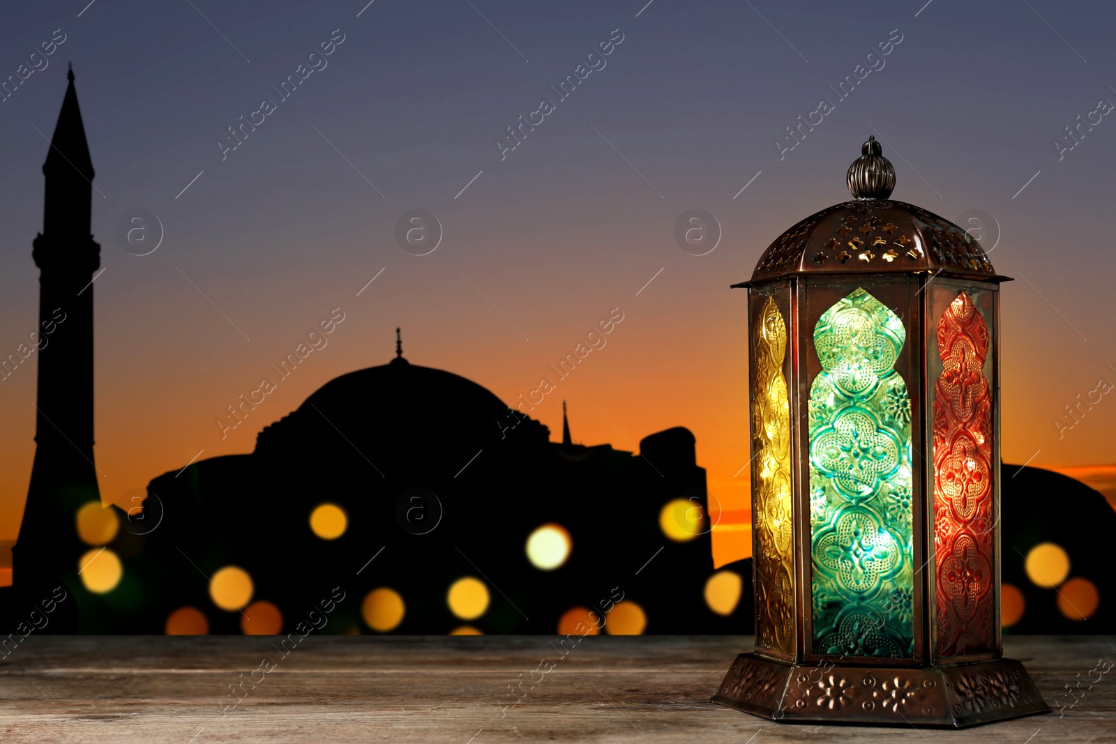 Image of Decorative Arabic lantern on wooden surface and silhouette of mosque at sunset on background, space for text
