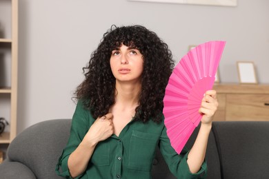 Photo of Young woman waving pink hand fan to cool herself on sofa at home