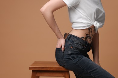 Woman with tattoo on belly sitting on stool against beige background, closeup