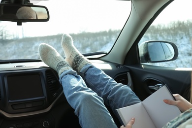 Young woman in warm socks holding her legs on car dashboard while reading book. Cozy atmosphere