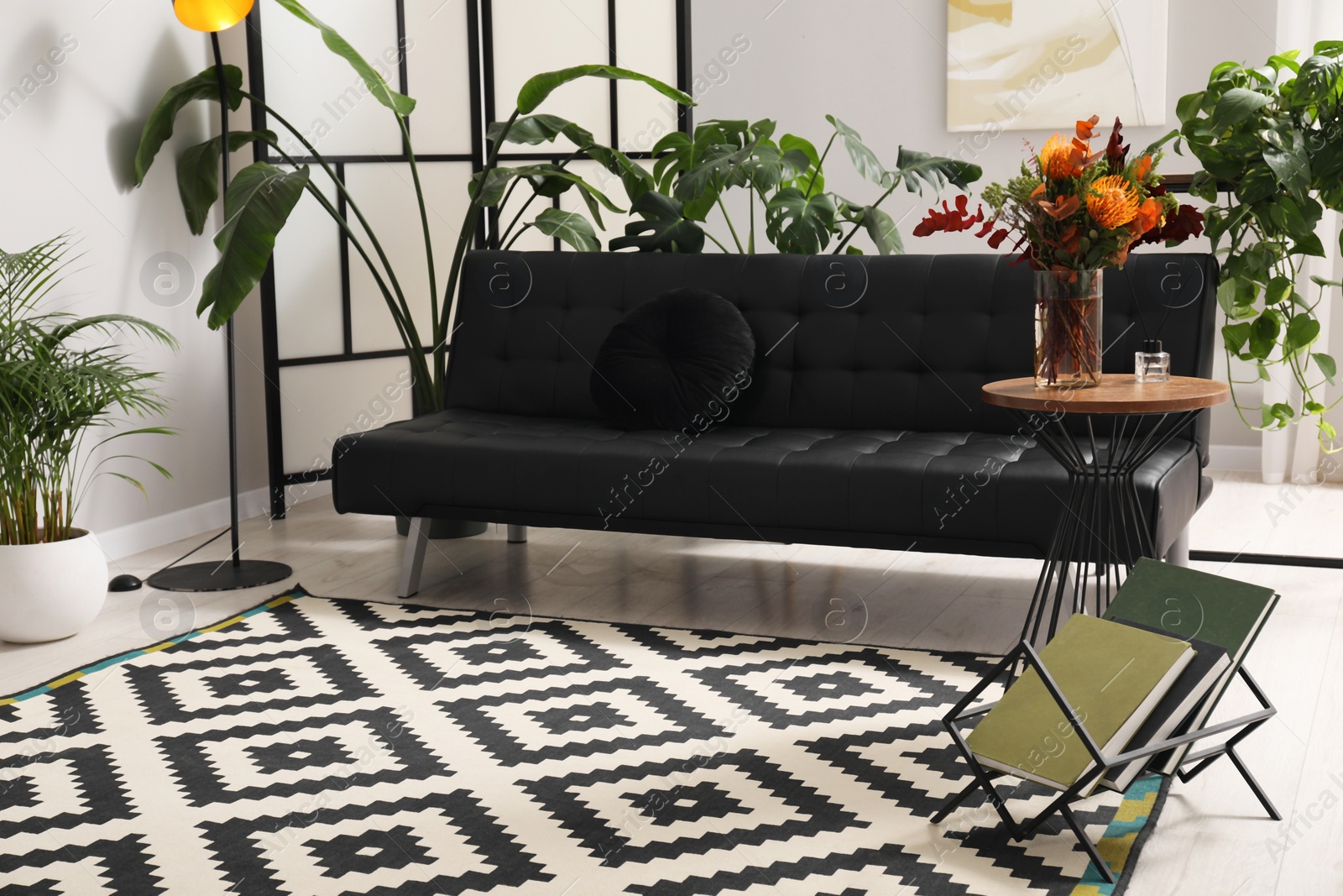 Photo of Vase with bouquet of beautiful leucospermum flowers on side table near black sofa and houseplants in room