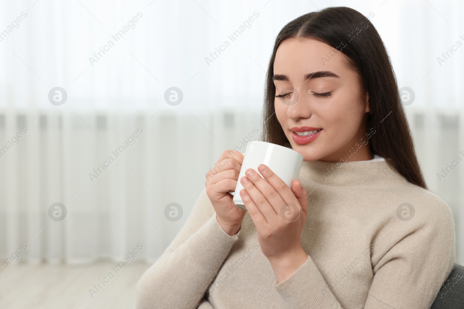 Photo of Beautiful young woman holding white ceramic mug at home, space for text