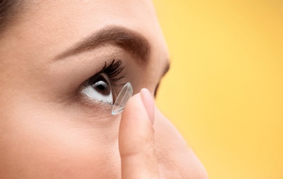 Young woman putting contact lens in her eye on color background, closeup