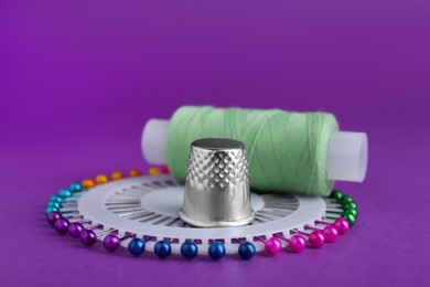 Photo of Thimble, pins and spool of light green sewing thread on purple background, closeup