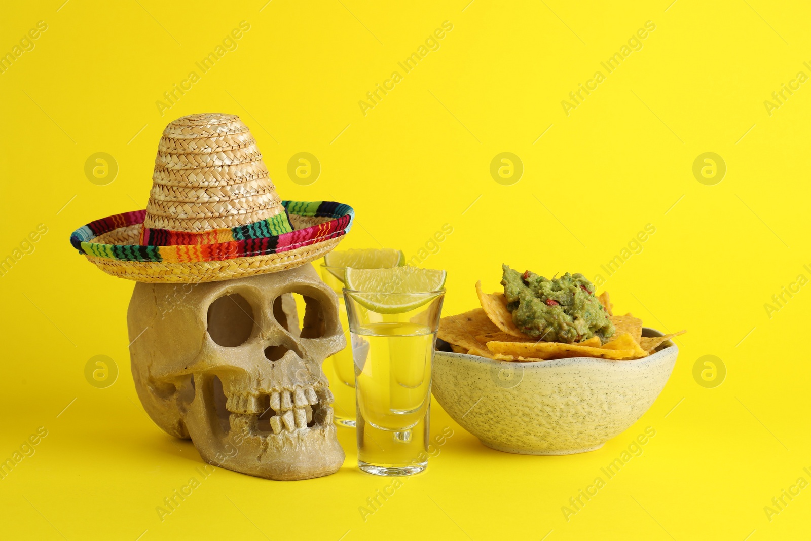 Photo of Mexican sombrero hat, human scull, tequila, nachos chips and guacamole in bowl on yellow background