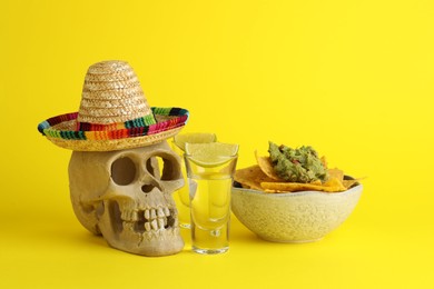 Mexican sombrero hat, human scull, tequila, nachos chips and guacamole in bowl on yellow background