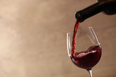 Pouring red wine into glass from bottle against blurred beige background, closeup. Space for text