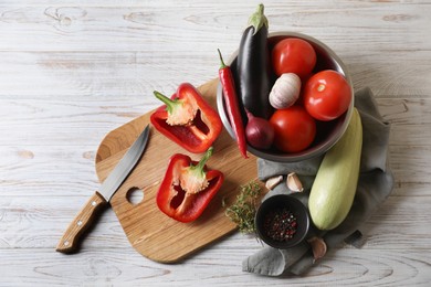 Photo of Cooking delicious ratatouille. Fresh ripe vegetables, knife and bowl on white wooden table, flat lay