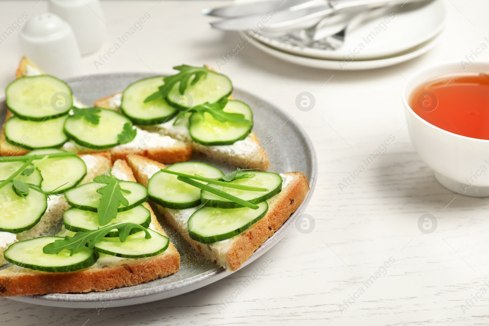 Photo of Plate with traditional English cucumber sandwiches and cup of tea on white wooden table