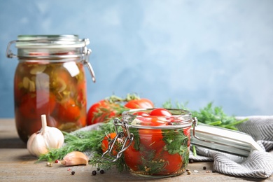 Pickled tomatoes in glass jars and products on wooden table against blue background, space for text