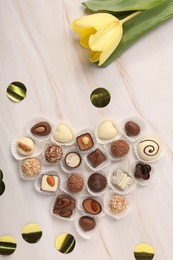 Heart made with delicious chocolate candies and beautiful tulip on beige marble table, flat lay
