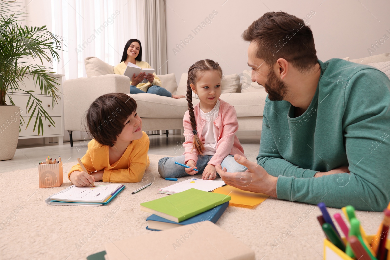 Photo of Father playing with his children while mother reading book on sofa in living room