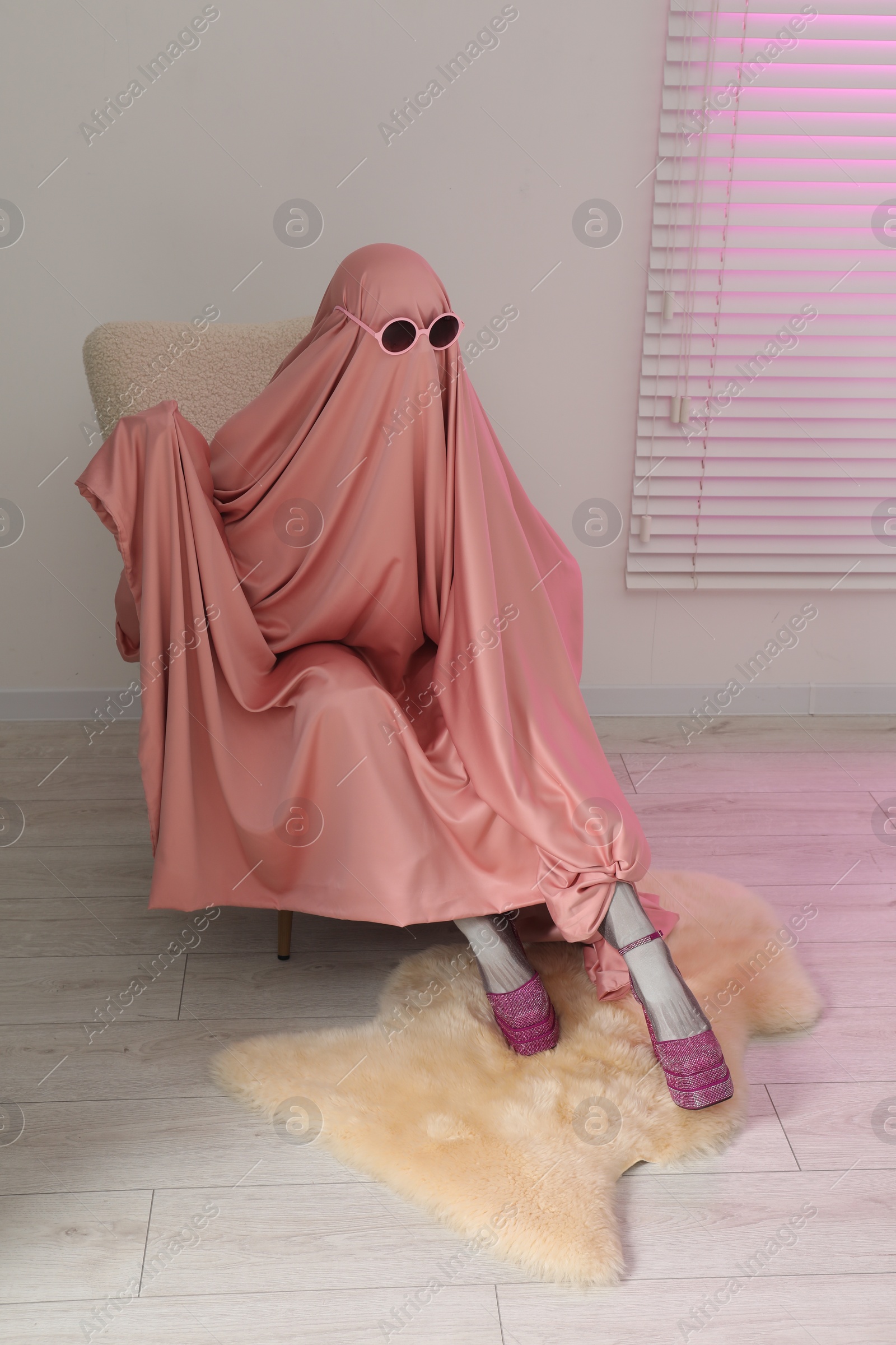 Photo of Glamorous ghost. Woman in pink sheet and high heel shoes on armchair indoors