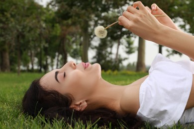 Beautiful young woman blowing dandelion while lying on green grass in park. Allergy free concept