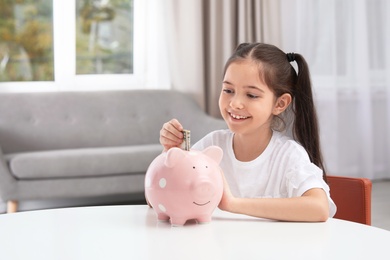 Photo of Little girl putting money into piggy bank at table indoors