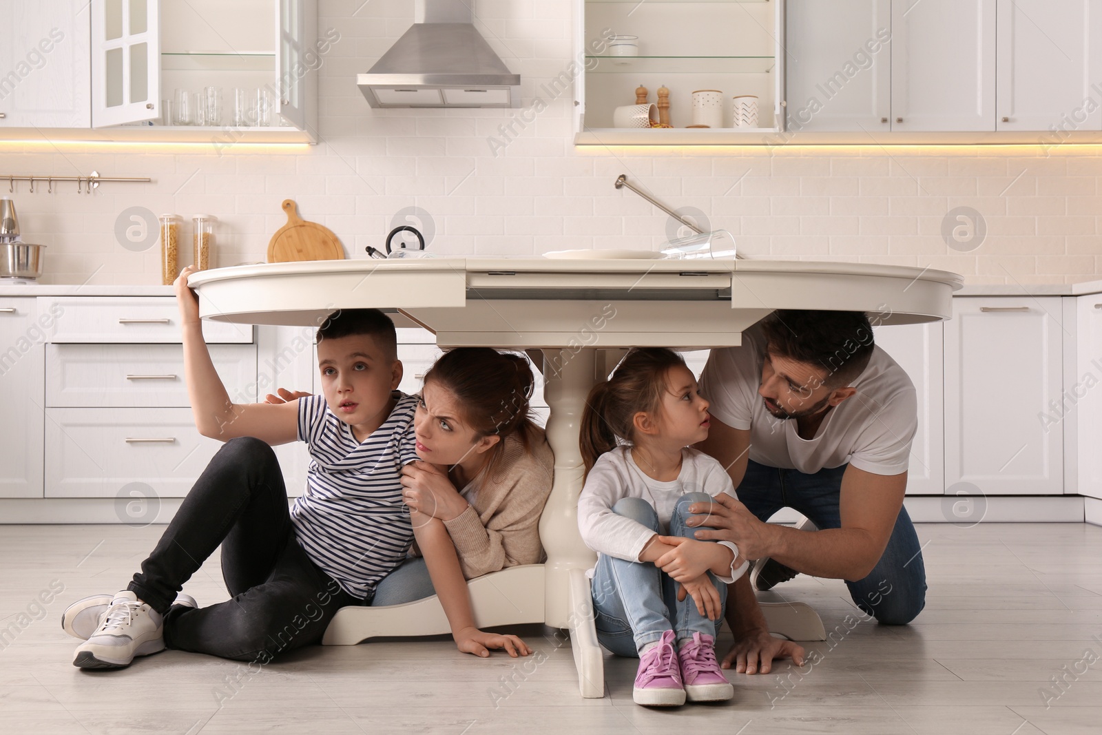 Photo of Scared parents with their children hiding under table in kitchen during earthquake