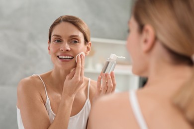 Photo of Beautiful woman removing makeup with cotton pad near mirror indoors