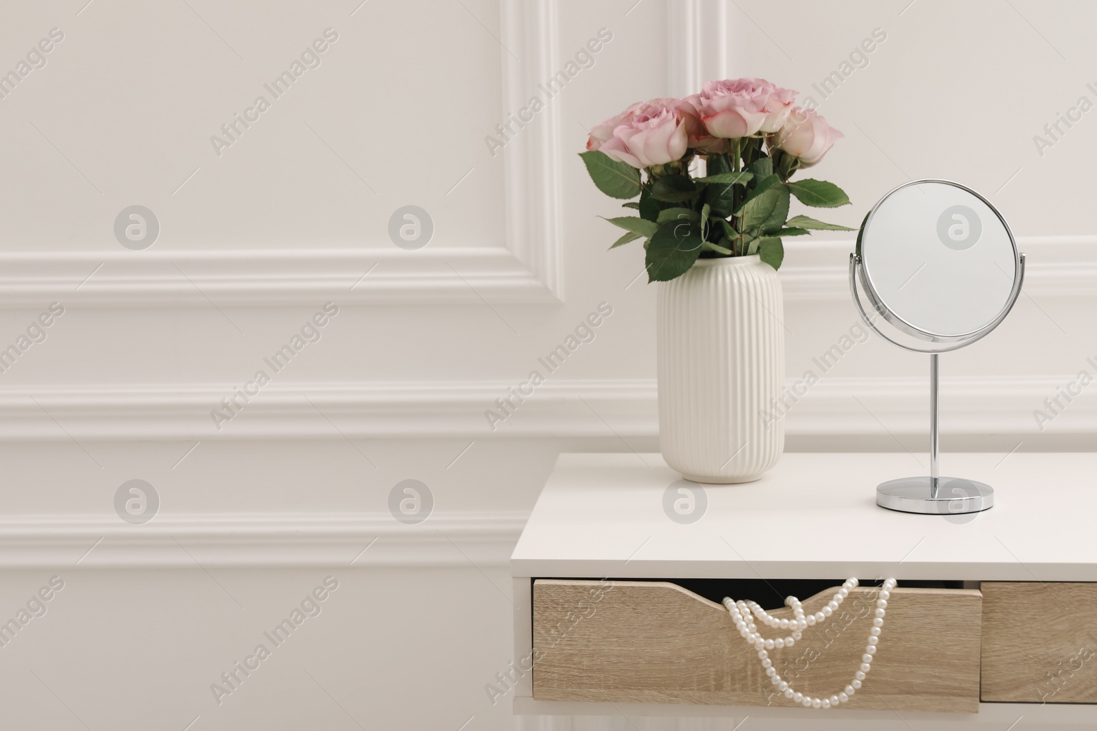 Photo of Mirror and vase with pink roses on white dressing table in makeup room, space for text