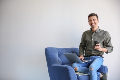 Young blogger with laptop sitting on armchair against light wall