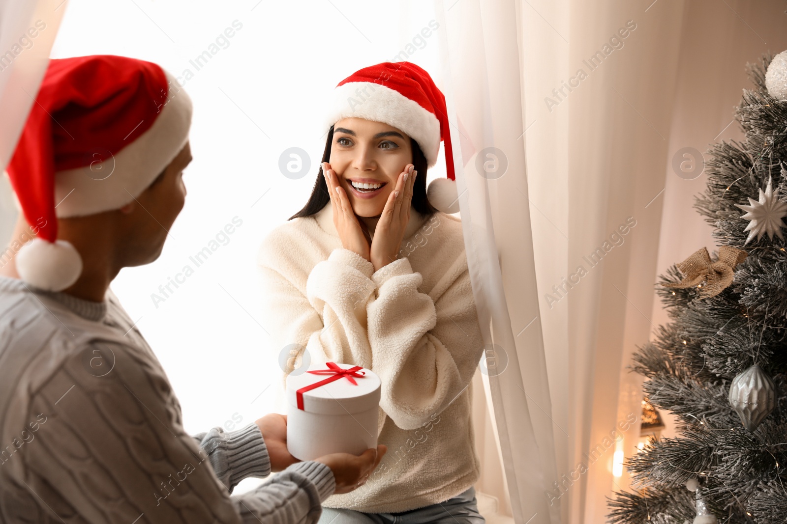 Photo of Boyfriend giving gift box to his girlfriend near Christmas tree