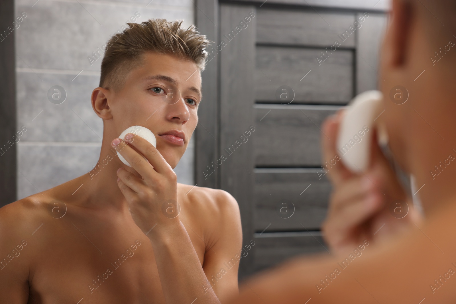 Photo of Young man washing his face with sponge near mirror in bathroom