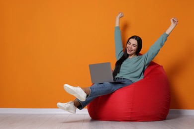 Photo of Cheerful woman with laptop sitting on beanbag chair near orange wall
