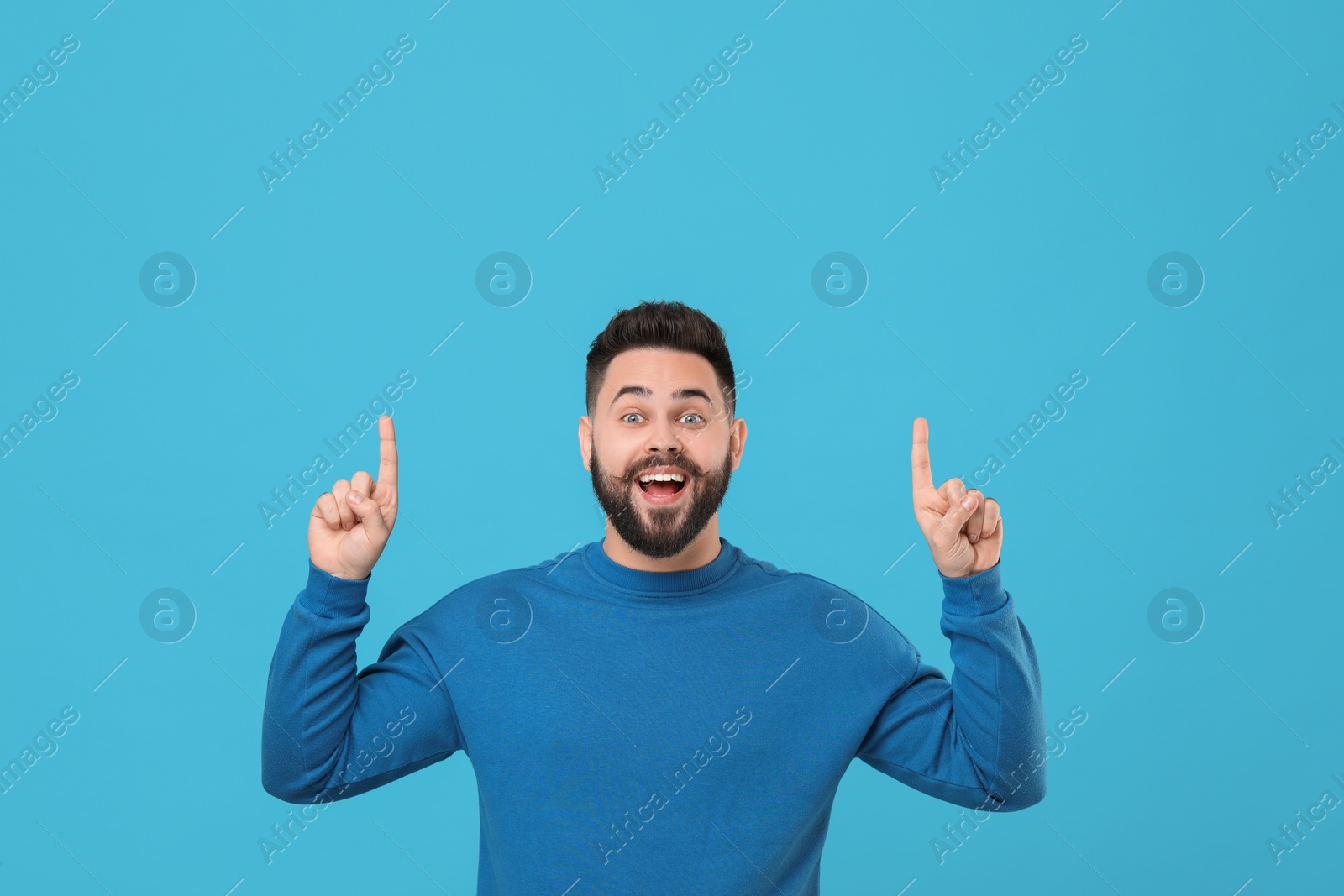 Photo of Happy young man with mustache pointing at something on light blue background. Space for text