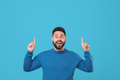 Happy young man with mustache pointing at something on light blue background. Space for text