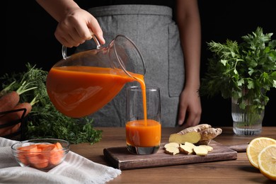 Woman pouring carrot juice from jug into glass at wooden table, closeup