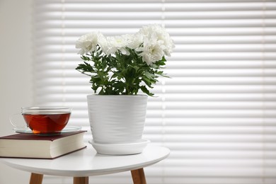 Beautiful chrysanthemum plant in flower pot, cup of tea and book on white table indoors, space for text