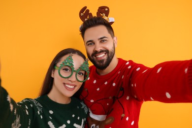 Photo of Happy young couple in Christmas sweaters, reindeer headband and party glasses taking selfie on orange background