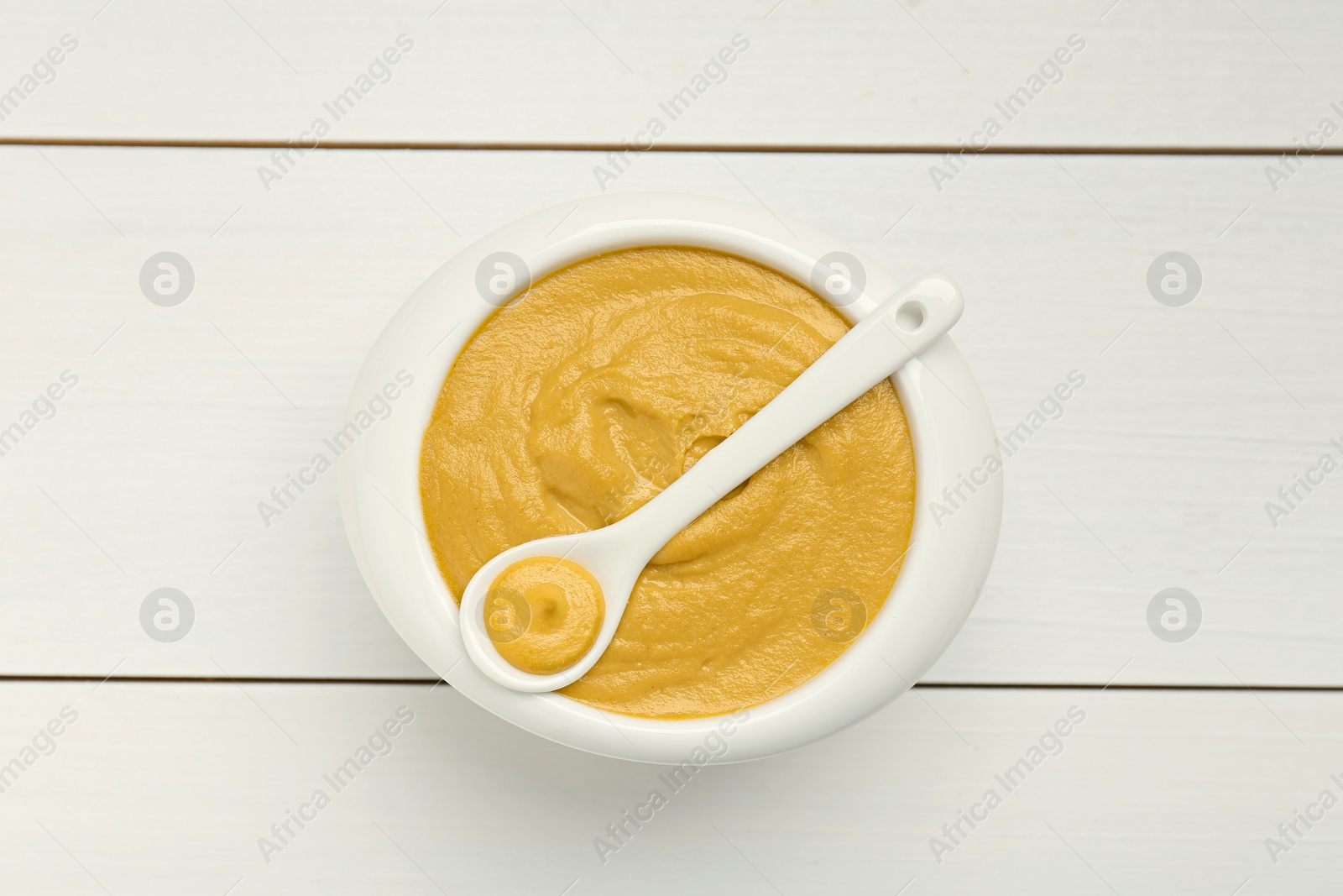 Photo of Spoon and bowl of tasty mustard sauce on white wooden table, top view