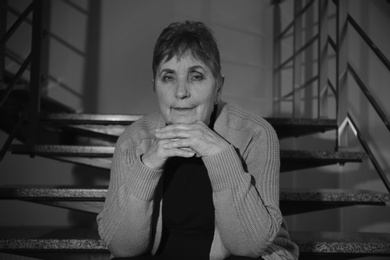 Photo of Poor senior woman sitting on stairs indoors. Black and white effect