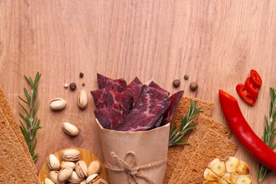Photo of Paper bag with pieces of delicious beef jerky and different spices on wooden table, flat lay. Space for text