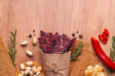 Paper bag with pieces of delicious beef jerky and different spices on wooden table, flat lay. Space for text