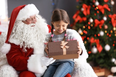 Photo of Little girl with gift box sitting on authentic Santa Claus' lap indoors