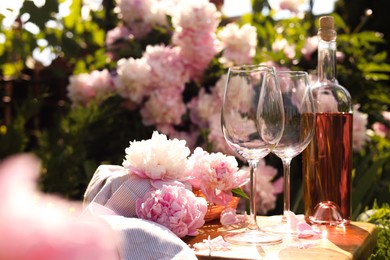 Bottle of rose wine and glasses with beautiful peonies on wooden table in garden