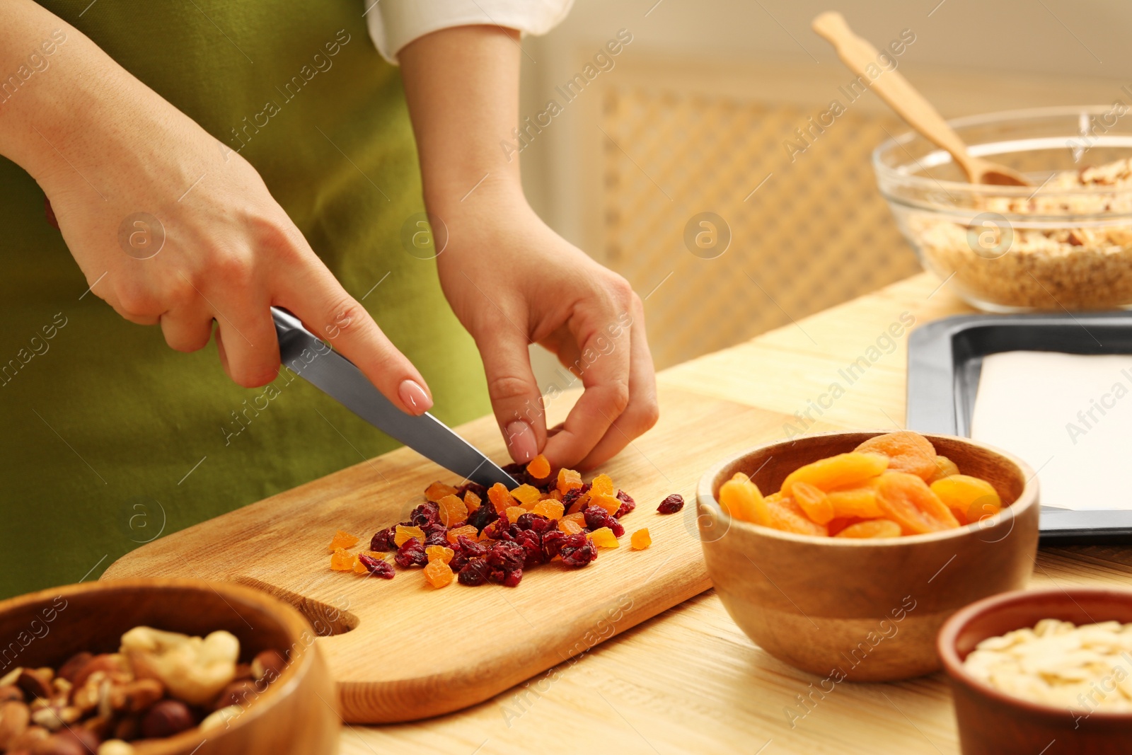 Photo of Making granola. Woman cutting dried apricots and cherries at table in kitchen, closeup