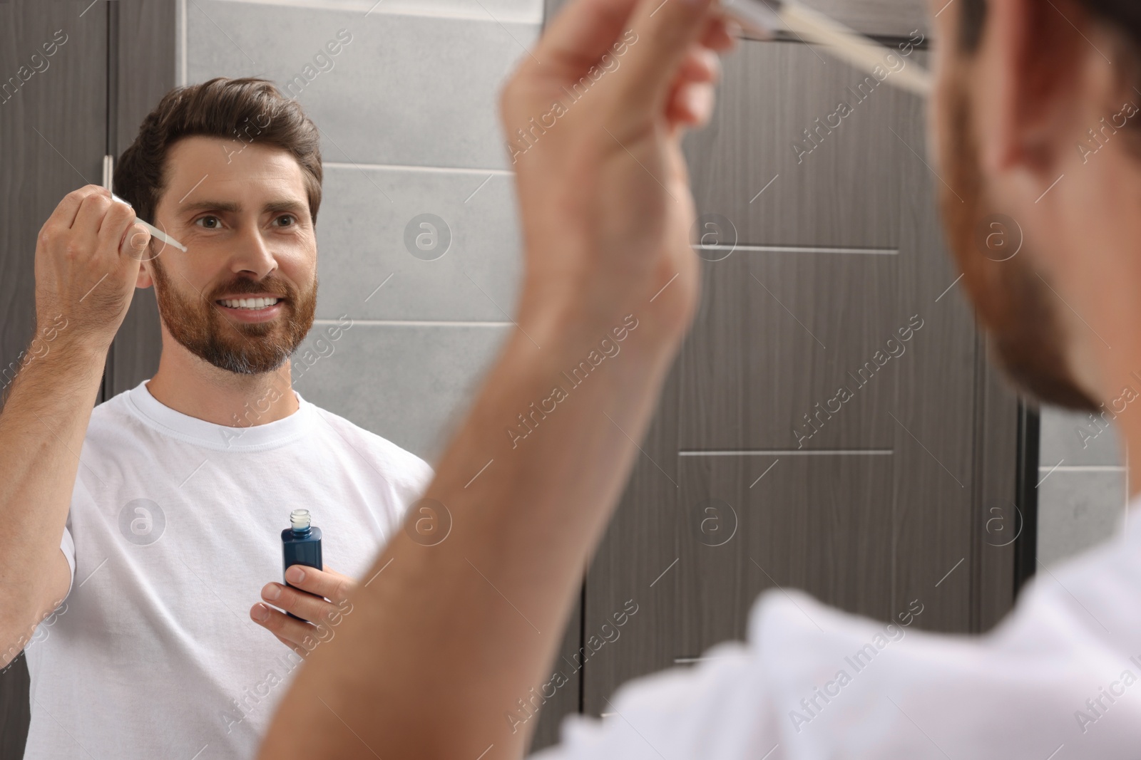 Photo of Handsome man applying oil on beard in bathroom