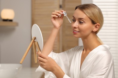 Beautiful woman applying cosmetic serum onto her face in front of mirror in bathroom