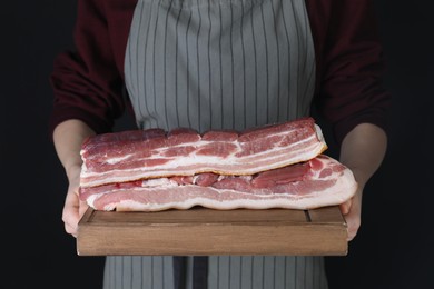 Woman holding wooden board with pieces of raw pork belly on black background, closeup