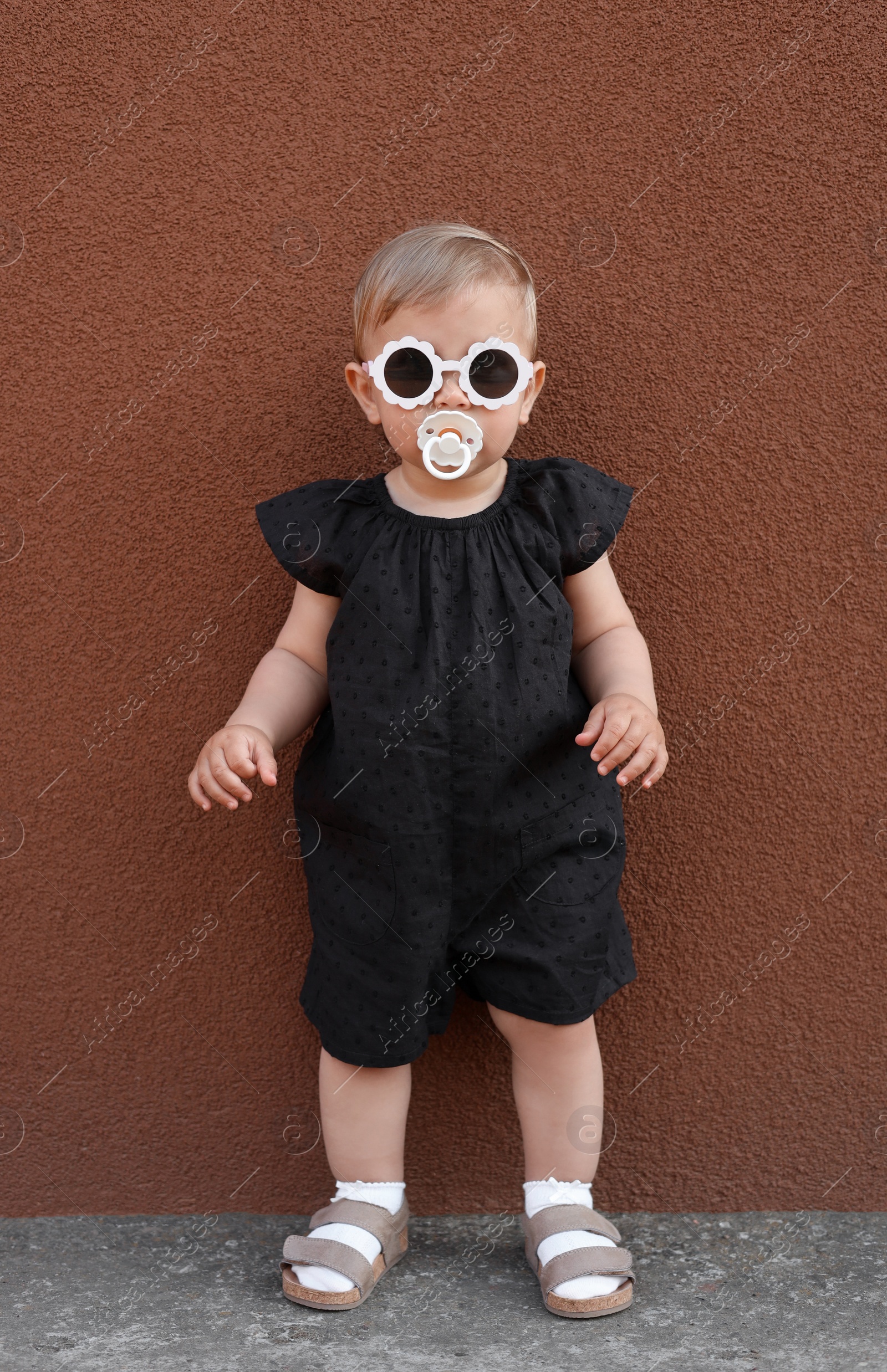 Photo of Cute little girl wearing stylish clothes with sunglasses and pacifier near brown wall outdoors