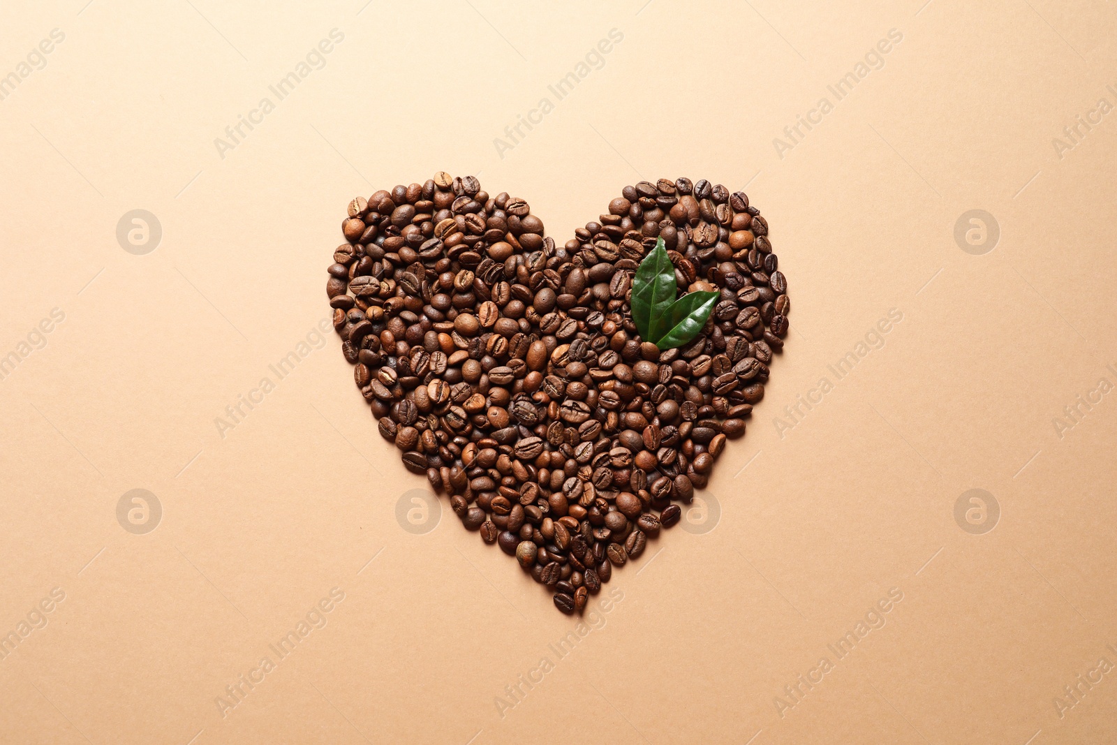 Photo of Heart shaped pile of coffee beans and fresh green leaves on light orange background, top view